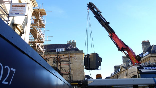 A substation is dangling from a crane arm above the hoarding at Bath Assembly Rooms and a wintery cold blue sky day.